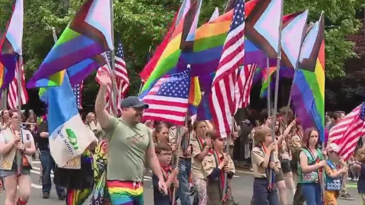Parade on a street with marchers carrying rainbow Pride flags and U.S. flags, celebrating together.