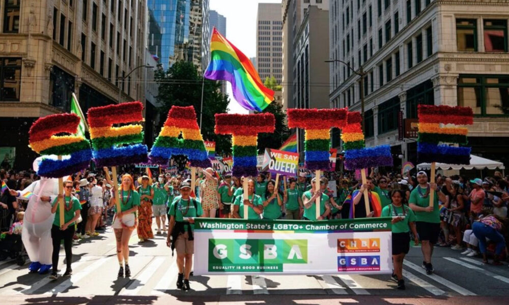 Pride parade on a city street with a large rainbow-letter float and a banner for LGBTQ+ groups marching together.