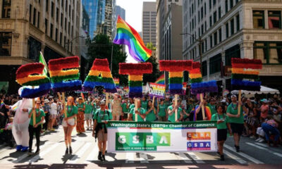 Pride parade on a city street with a large rainbow-letter float and a banner for LGBTQ+ groups marching together.