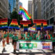 Pride parade on a city street with a large rainbow-letter float and a banner for LGBTQ+ groups marching together.