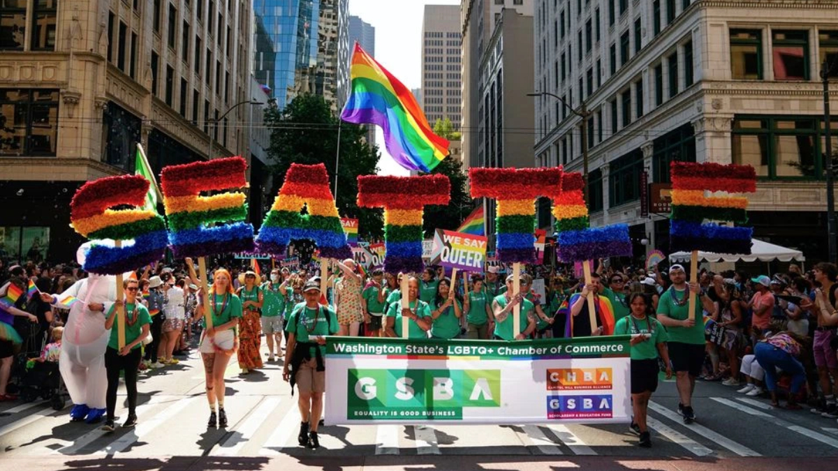 Pride parade on a city street with a large rainbow-letter float and a banner for LGBTQ+ groups marching together.