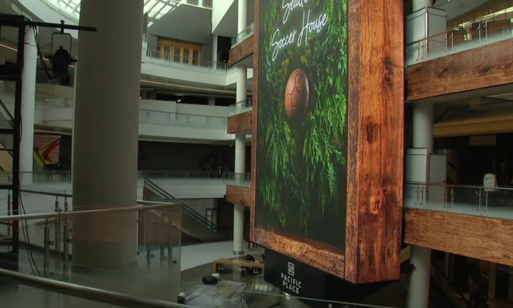 Indoor mall atrium with a tall hanging digital banner showing a soccer ball among greenery, Pacific Place logo at bottom surface area visible.