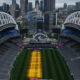 Aerial view of a large stadium with a green field and a yellow central court, city skyline in the background.” but we must remove smart quotes.