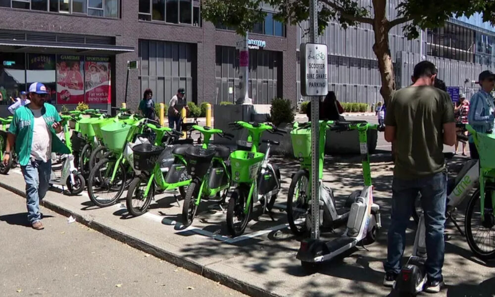 Row of lime-green bike-share bicycles parked along a city sidewalk with pedestrians nearby on a sunny day.