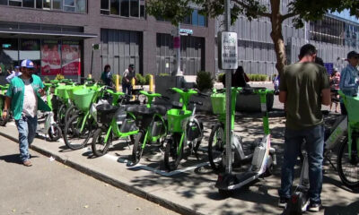 Row of lime-green bike-share bicycles parked along a city sidewalk with pedestrians nearby on a sunny day.