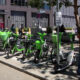 Row of lime-green bike-share bicycles parked along a city sidewalk with pedestrians nearby on a sunny day.