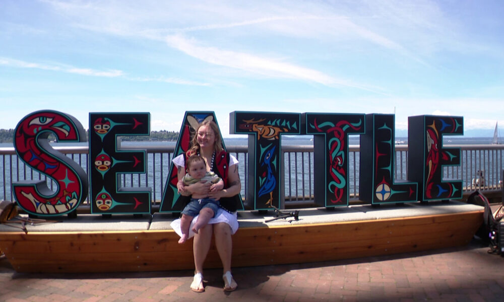 Woman sitting on a wooden platform holding a baby in front of a colorful Seattle sign by the water under a blue sky.