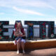 Woman sitting on a wooden platform holding a baby in front of a colorful Seattle sign by the water under a blue sky.