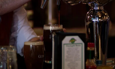 Bartender pours dark stout from a tap into a pint glass at a bar, with another Guinness glass nearby.