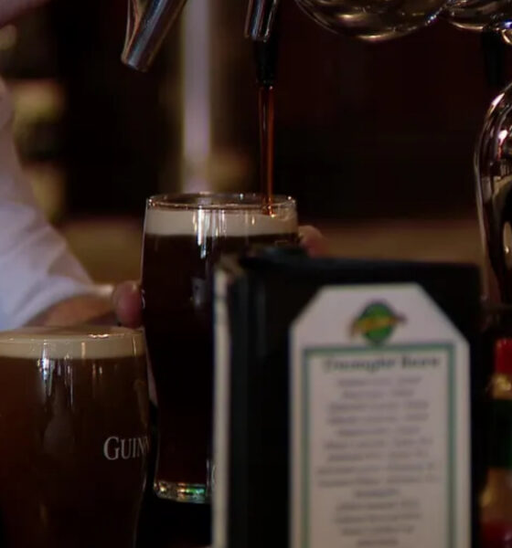 Bartender pours dark stout from a tap into a pint glass at a bar, with another Guinness glass nearby.