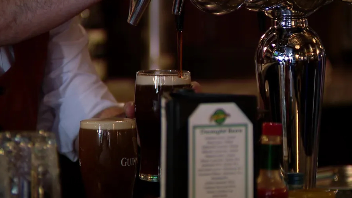 Bartender pours dark stout from a tap into a pint glass at a bar, with another Guinness glass nearby.