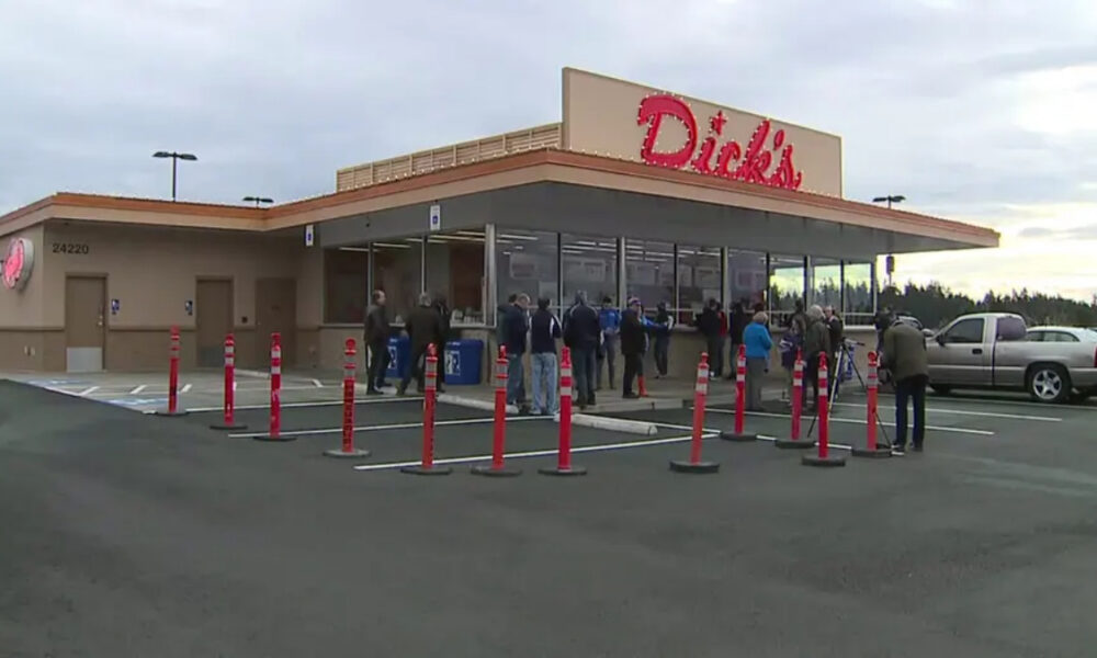People line up outside a Dick's store, waiting near the entrance with orange cones guiding the line in the parking lot.