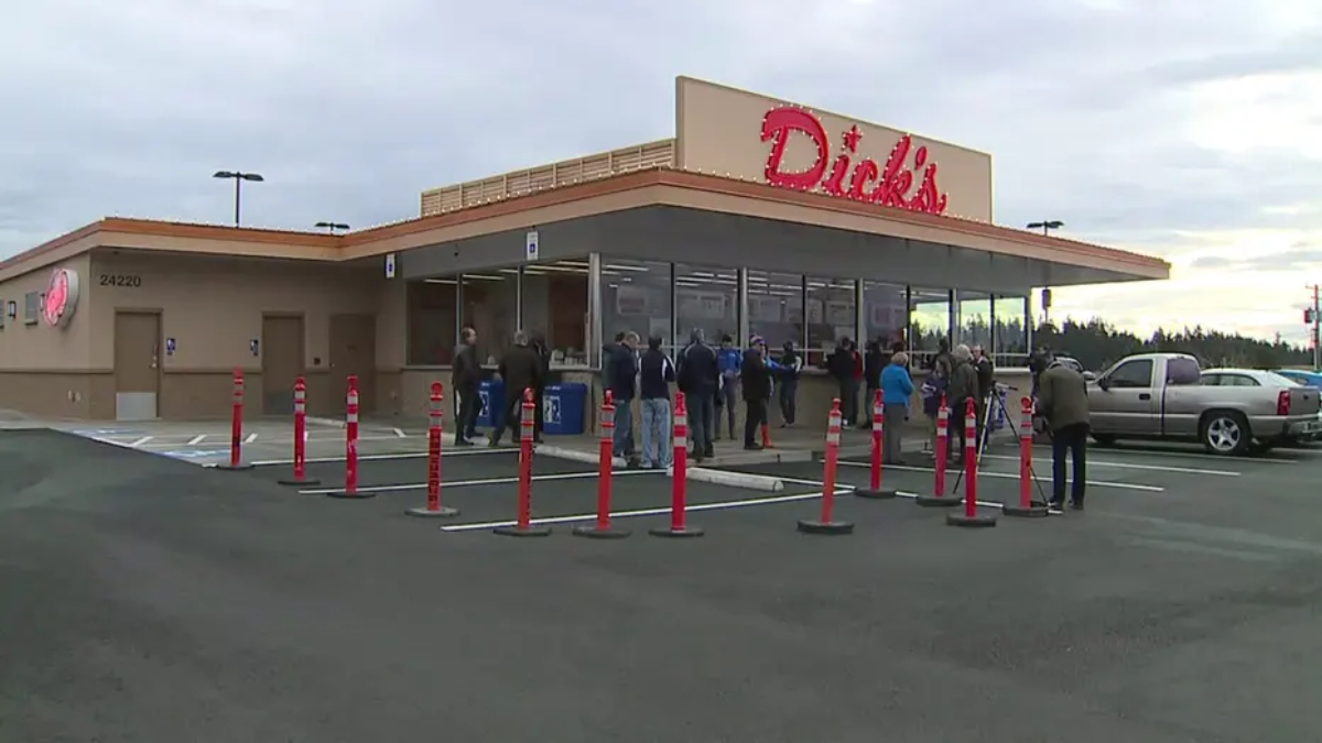 People line up outside a Dick's store, waiting near the entrance with orange cones guiding the line in the parking lot.