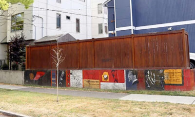 Low wall painted with colorful panels and a small, leafless tree in front, with a tall brown fence and modern buildings behind.