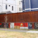 Low wall painted with colorful panels and a small, leafless tree in front, with a tall brown fence and modern buildings behind.