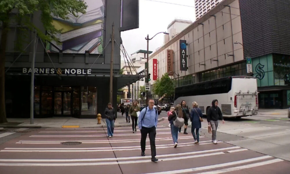Pedestrians cross a city street at a wide crosswalk in front of Barnes & Noble; a white bus is on the right side.