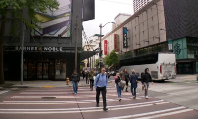 Pedestrians cross a city street at a wide crosswalk in front of Barnes & Noble; a white bus is on the right side.