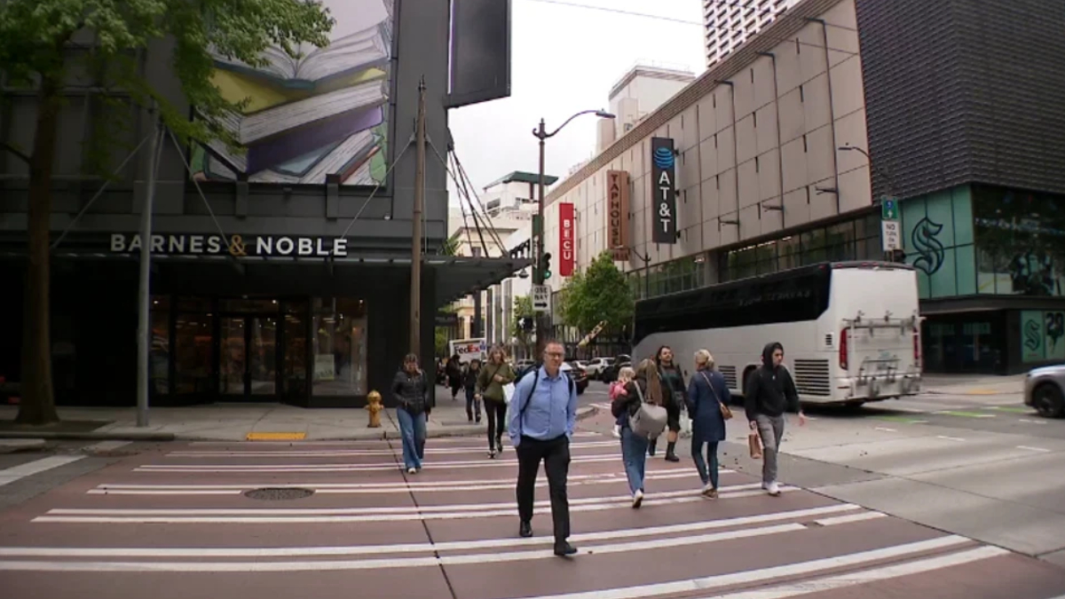 Pedestrians cross a city street at a wide crosswalk in front of Barnes & Noble; a white bus is on the right side.