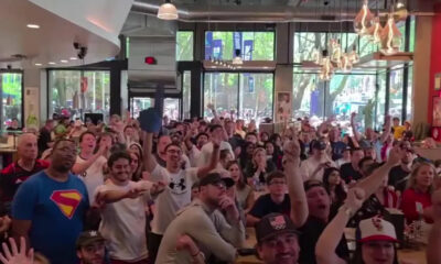Large crowd of people inside a bright cafe watching an event, many with raised hands and smiles.