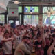 Large crowd of people inside a bright cafe watching an event, many with raised hands and smiles.