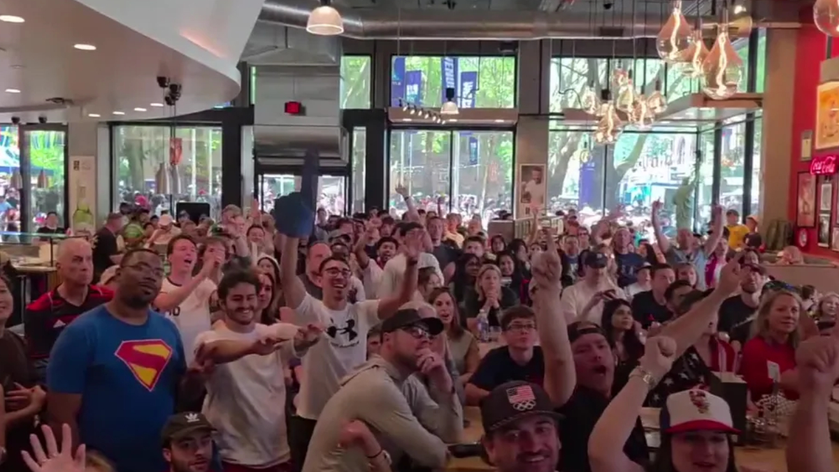 Large crowd of people inside a bright cafe watching an event, many with raised hands and smiles.
