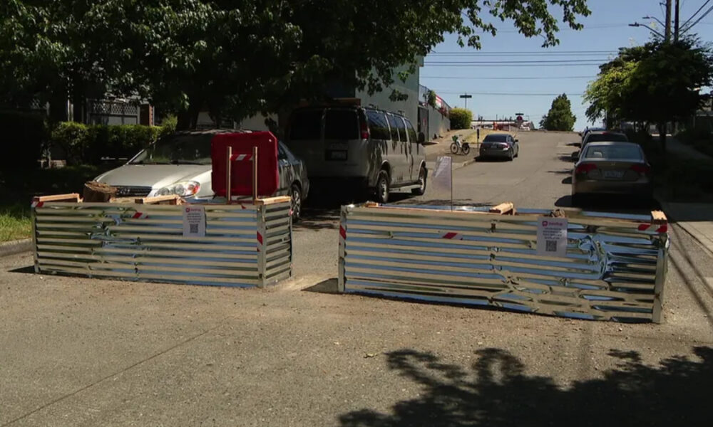 Two metal construction barricades block a sunlit residential street, with parked cars behind and a van nearby.