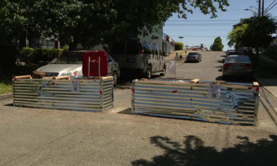 Two metal construction barricades block a sunlit residential street, with parked cars behind and a van nearby.