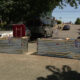 Two metal construction barricades block a sunlit residential street, with parked cars behind and a van nearby.