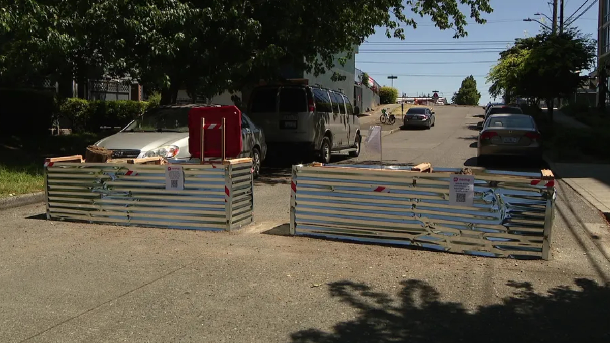 Two metal construction barricades block a sunlit residential street, with parked cars behind and a van nearby.