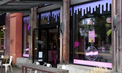 Exterior of a donut shop with pink Donuts & Coffee sign, wooden posts, and white outdoor chairs outside.