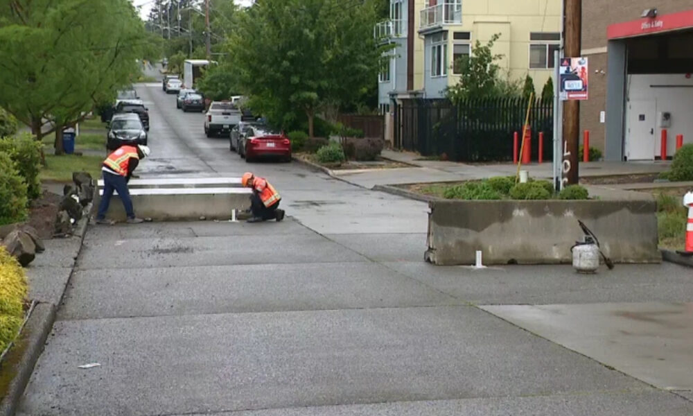 Two workers in orange safety vests repair a curb on a residential street, with barriers and equipment nearby.