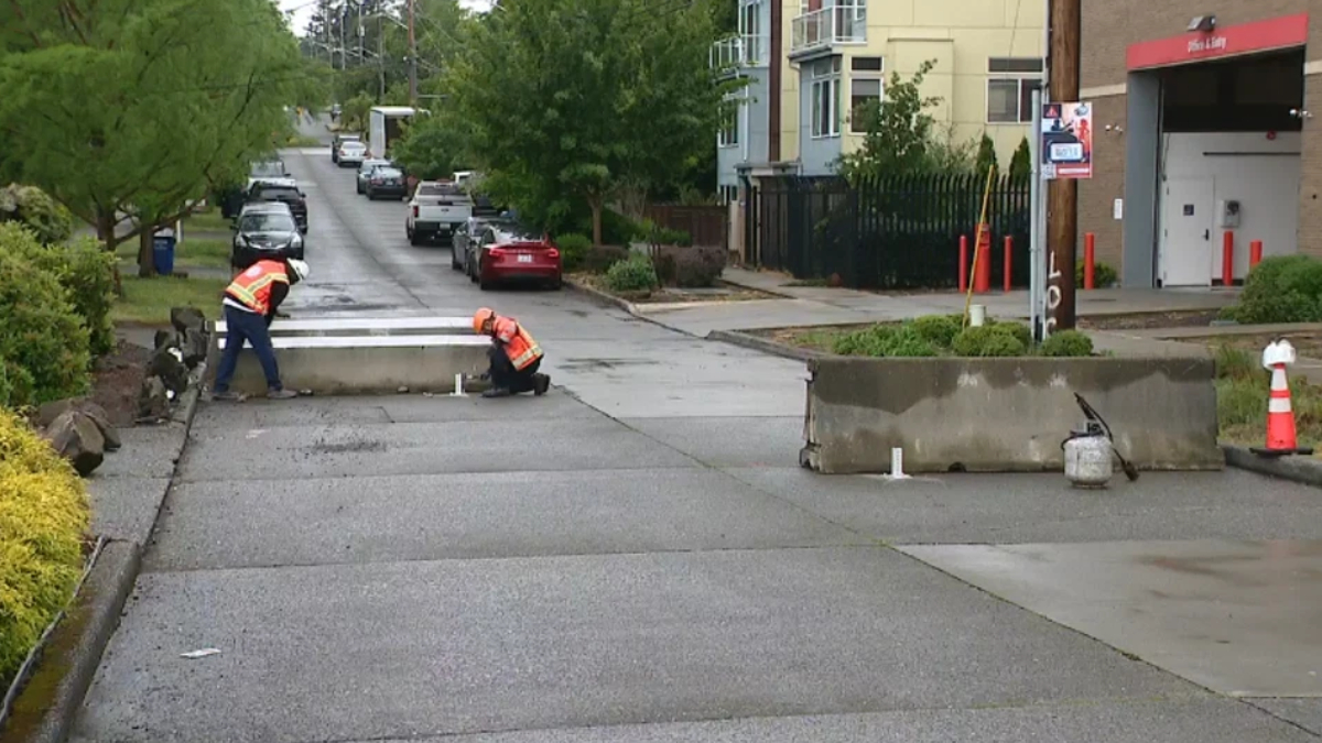 Two workers in orange safety vests repair a curb on a residential street, with barriers and equipment nearby.