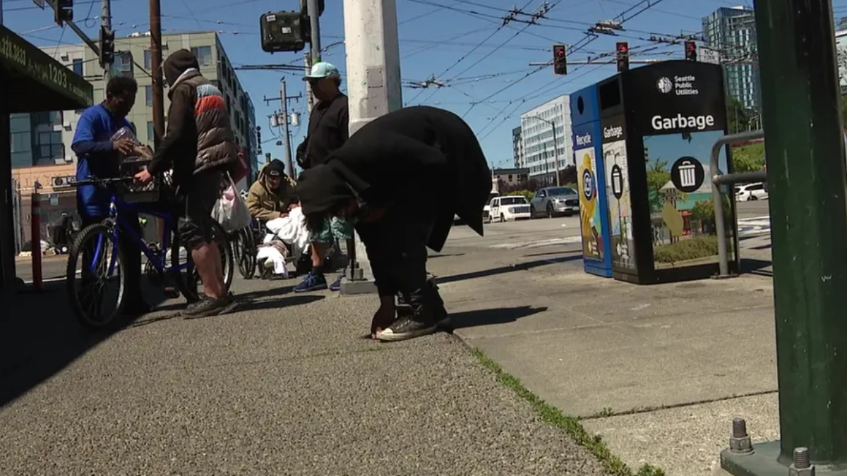 Urban street scene with people and bicycles gathered on the sidewalk, a person in black bends down near the curb while others chat nearby.