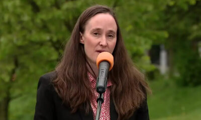 Woman with long brown hair wearing a black blazer speaks into an orange microphone outdoors with green trees in the background.