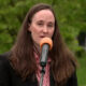 Woman with long brown hair wearing a black blazer speaks into an orange microphone outdoors with green trees in the background.