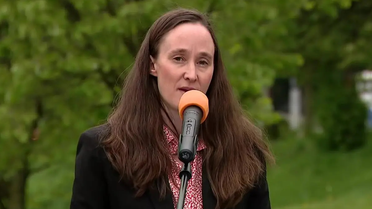 Woman with long brown hair wearing a black blazer speaks into an orange microphone outdoors with green trees in the background.