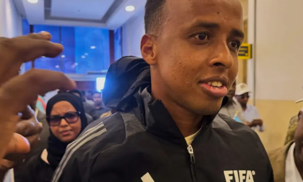 Man wearing a black FIFA jacket being surrounded by a crowd in a corridor, speaking or answering questions.
