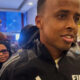 Man wearing a black FIFA jacket being surrounded by a crowd in a corridor, speaking or answering questions.