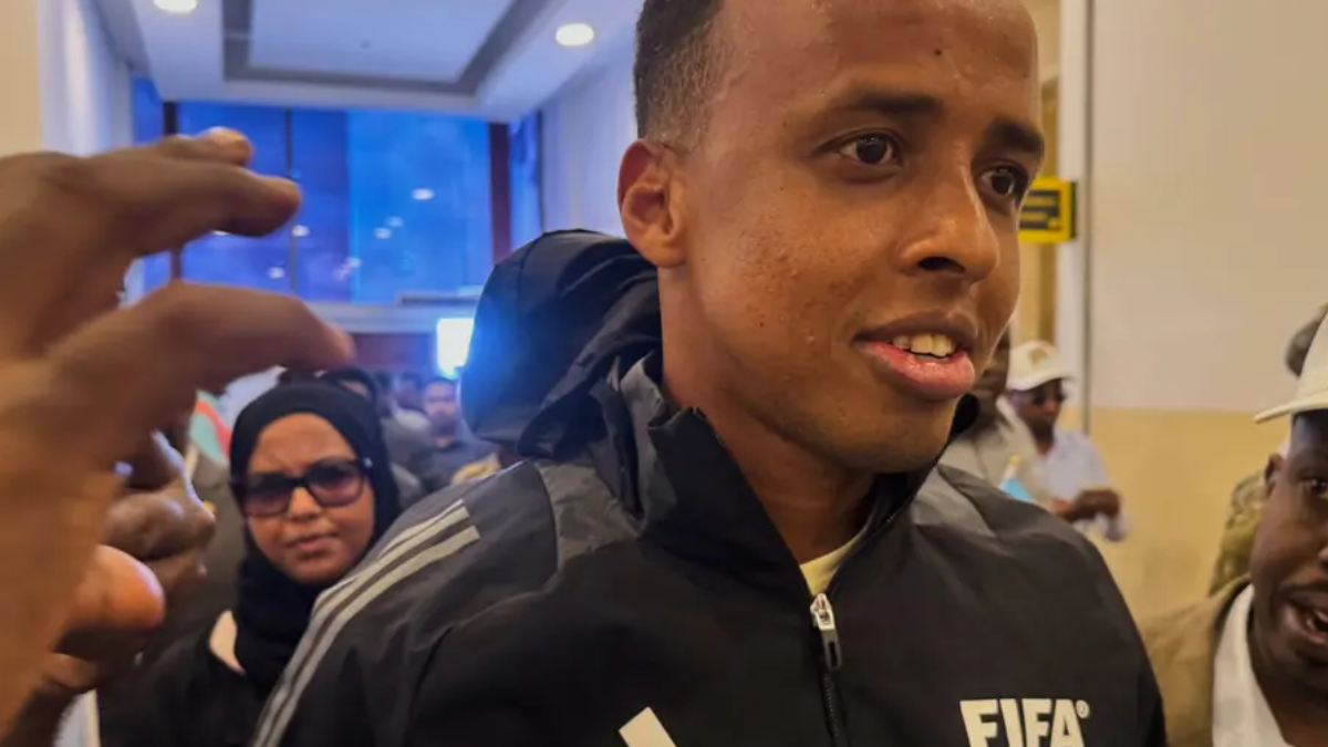 Man wearing a black FIFA jacket being surrounded by a crowd in a corridor, speaking or answering questions.