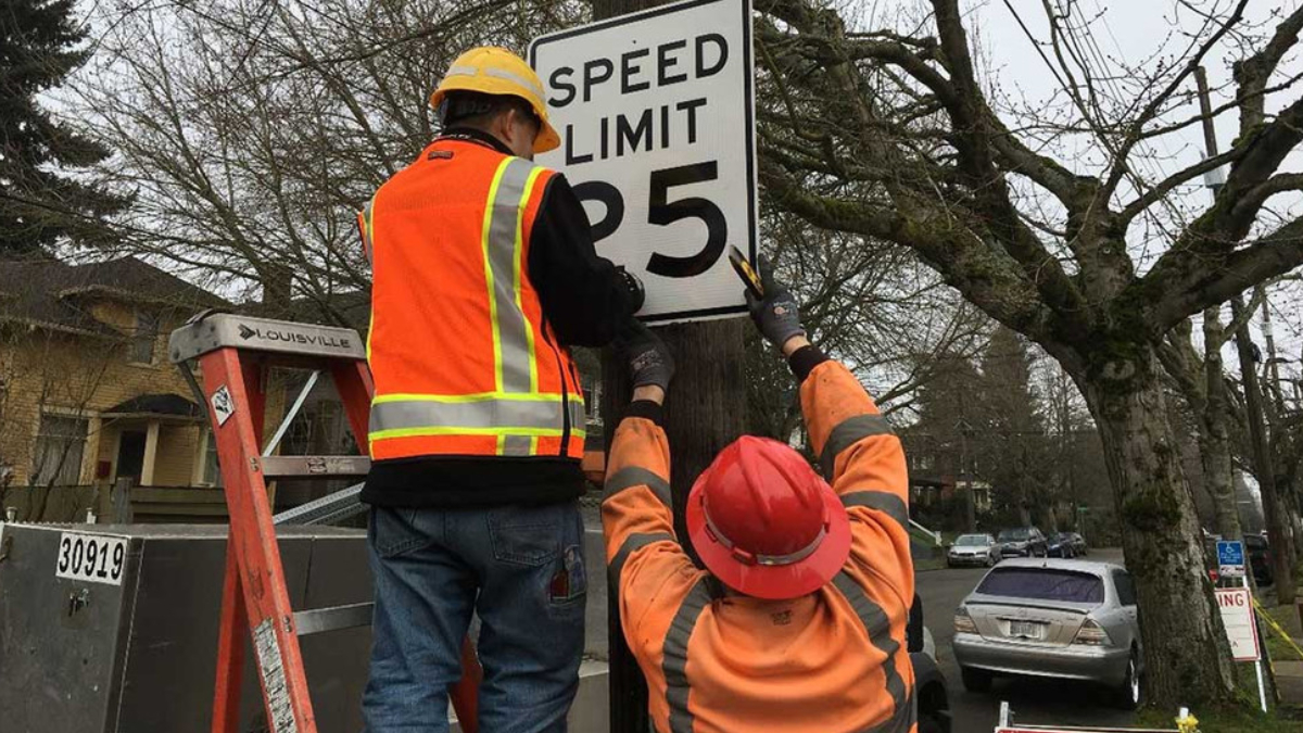 Two construction workers in high-visibility vests and hard hats mount a SPEED LIMIT 25 sign on a utility pole adjacent to a residential street.