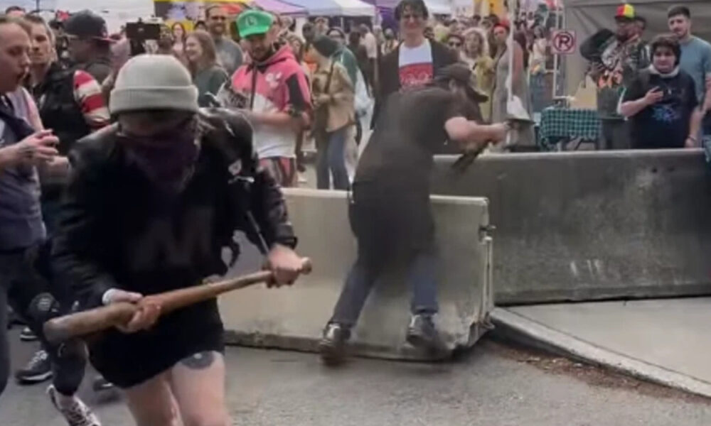 Man in a gray beanie and black clothing swings a wooden bat near a barricade at a crowded street event; onlookers surround the scene.