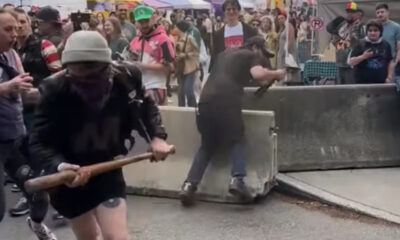 Man in a gray beanie and black clothing swings a wooden bat near a barricade at a crowded street event; onlookers surround the scene.