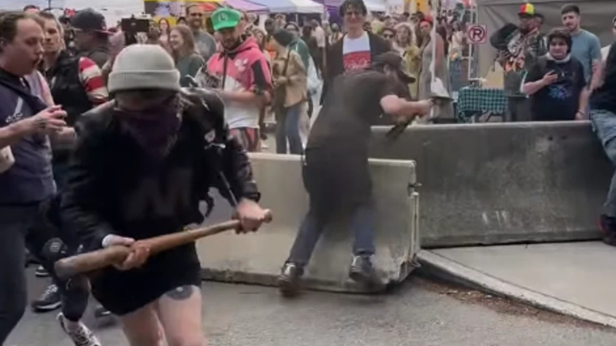Man in a gray beanie and black clothing swings a wooden bat near a barricade at a crowded street event; onlookers surround the scene.