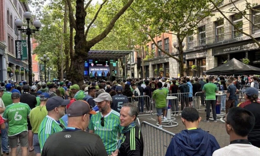 Crowd of fans in green jerseys gathered behind barriers for an outdoor viewing event on a city street with a stage and large screen in the background.