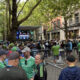 Crowd of fans in green jerseys gathered behind barriers for an outdoor viewing event on a city street with a stage and large screen in the background.