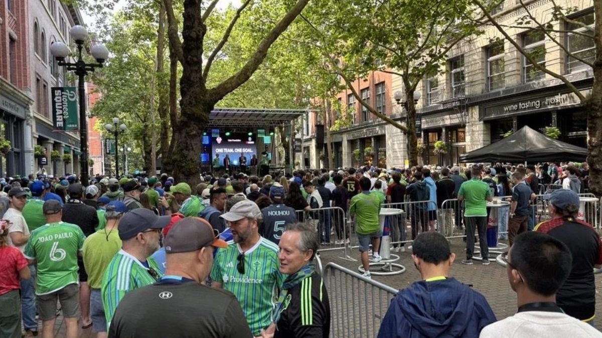 Crowd of fans in green jerseys gathered behind barriers for an outdoor viewing event on a city street with a stage and large screen in the background.