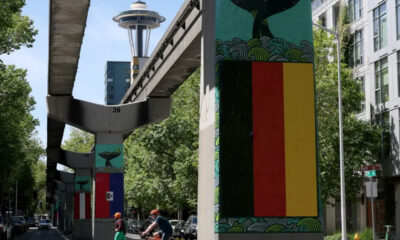 Urban street with an elevated rail overpass; mural-covered columns, including a pane resembling the German flag, and the Space Needle in the distance.
