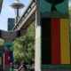 Urban street with an elevated rail overpass; mural-covered columns, including a pane resembling the German flag, and the Space Needle in the distance.