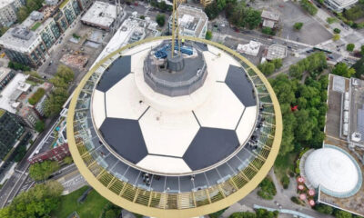 Aerial view of a circular observation tower with a ring walkway atop a city blocked by trees and buildings nearby.