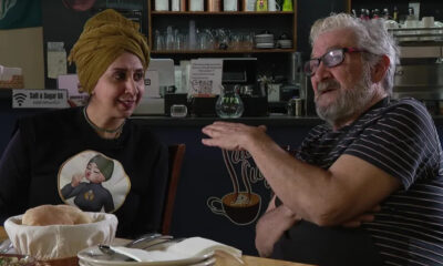 Two people sit at a cafe table and chat; a woman with a mustard head wrap smiles while a bearded man gestures with his hands.